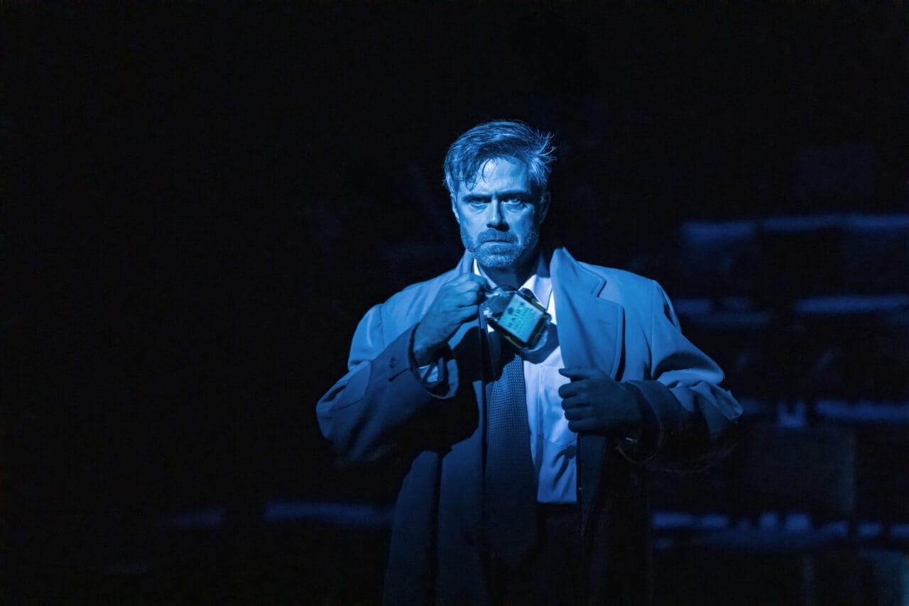 A man in a trench coat stands in dramatic blue lighting, holding a small radio to his mouth with a serious expression—like a scene straight from a John le Carré spy novel.