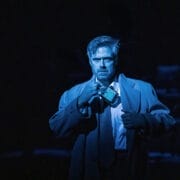 A man in a trench coat stands in dramatic blue lighting, holding a small radio to his mouth with a serious expression—like a scene straight from a John le Carré spy novel.