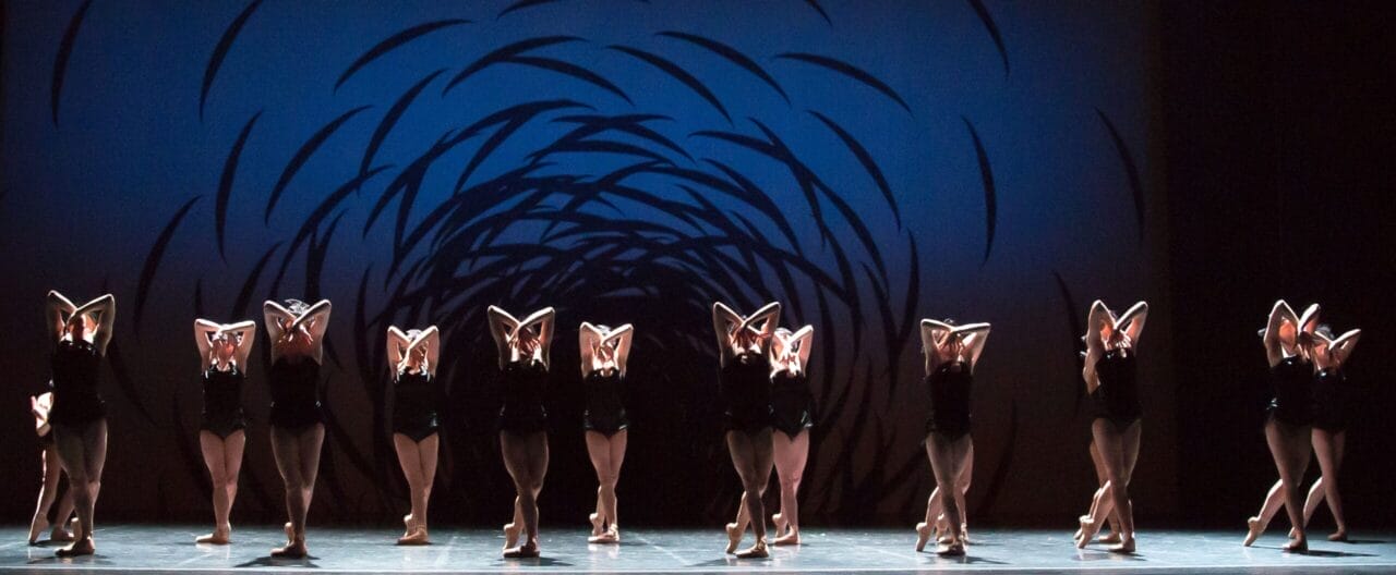 A group of dancers in black costumes perform on stage, standing in a line with arms raised and crossed, against a blue background with a swirling black pattern.