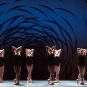 A group of dancers in black costumes perform on stage, standing in a line with arms raised and crossed, against a blue background with a swirling black pattern.