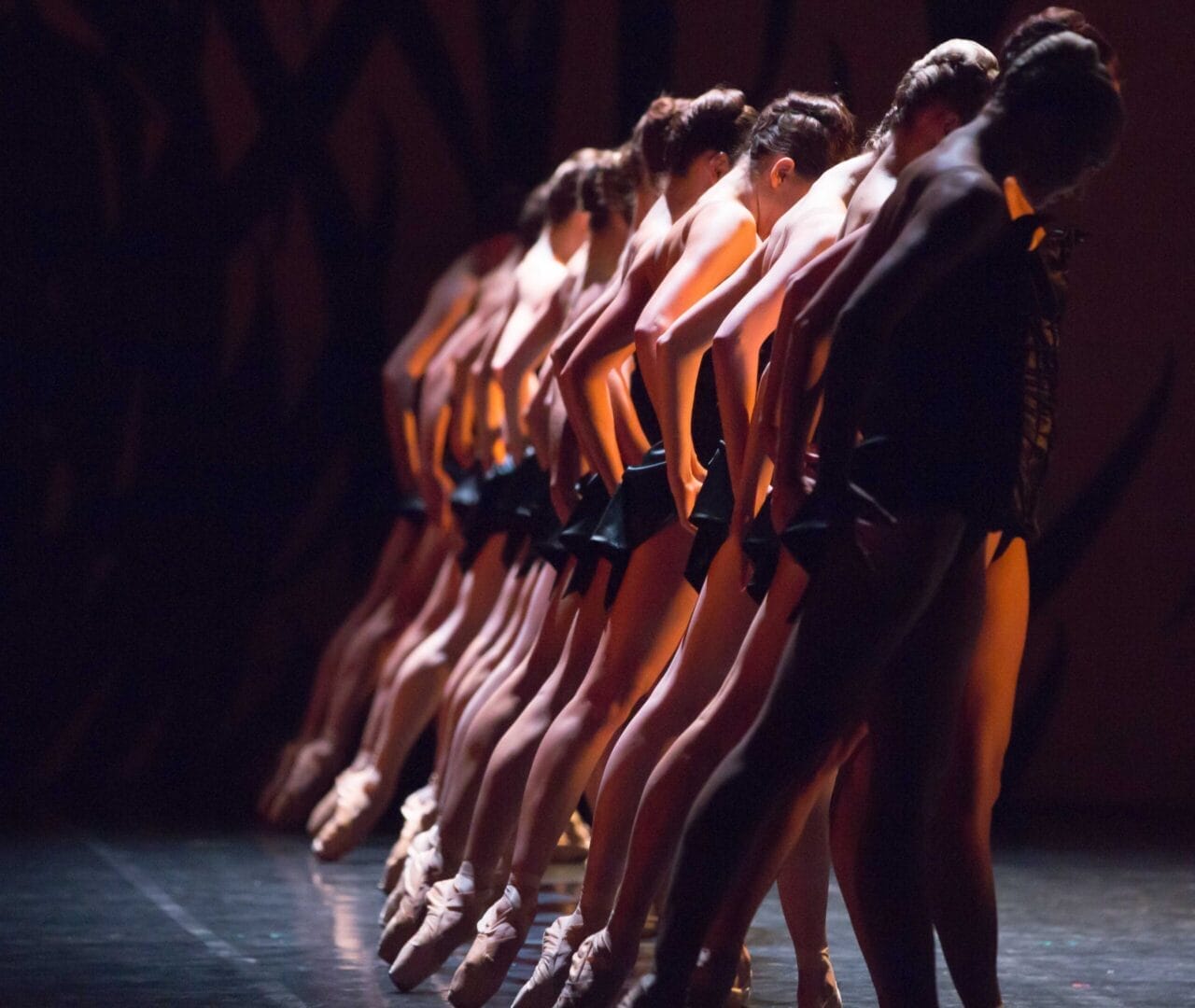 A line of ballet dancers in black costumes and pointe shoes perform a synchronized pose on stage, illuminated by stage lighting.