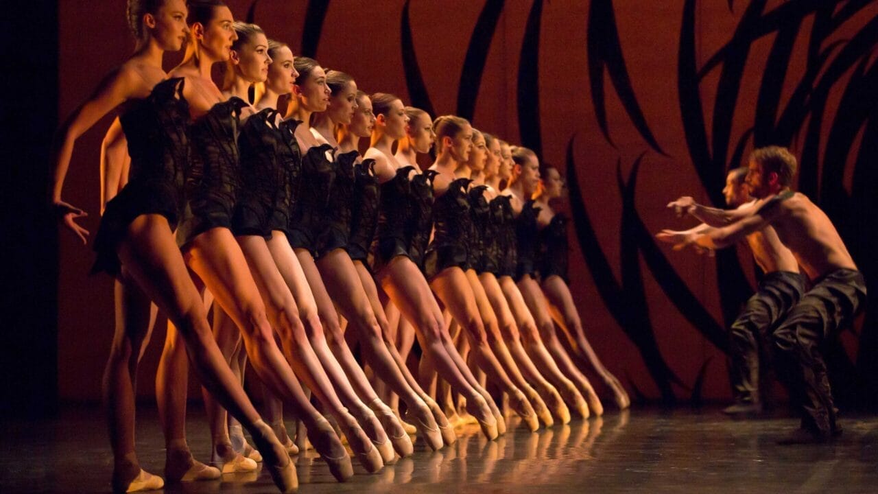 A line of ballerinas in black costumes performs on stage en pointe, while two male dancers face them with arms extended against an orange backdrop with black abstract designs.