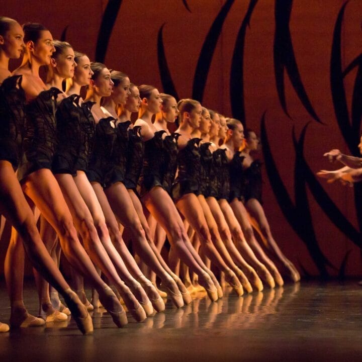 A line of ballerinas in black costumes performs on stage en pointe, while two male dancers face them with arms extended against an orange backdrop with black abstract designs.