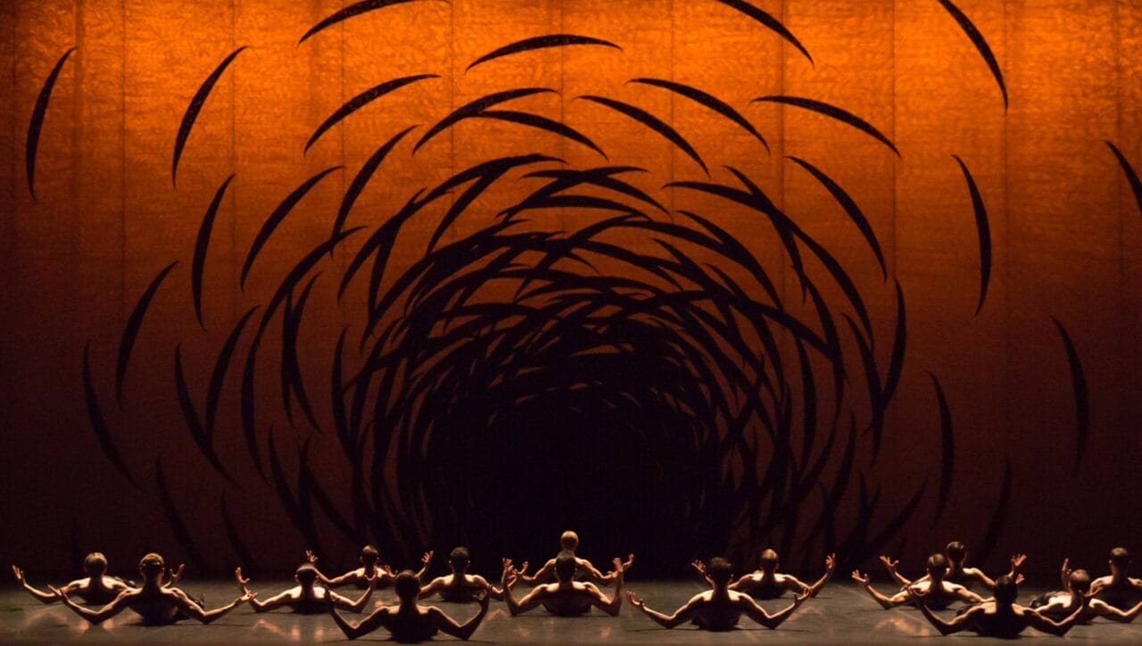 A group of dancers sit on stage facing an orange backdrop with swirling black patterns that create a tunnel-like visual effect.
