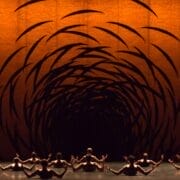 A group of dancers sit on stage facing an orange backdrop with swirling black patterns that create a tunnel-like visual effect.