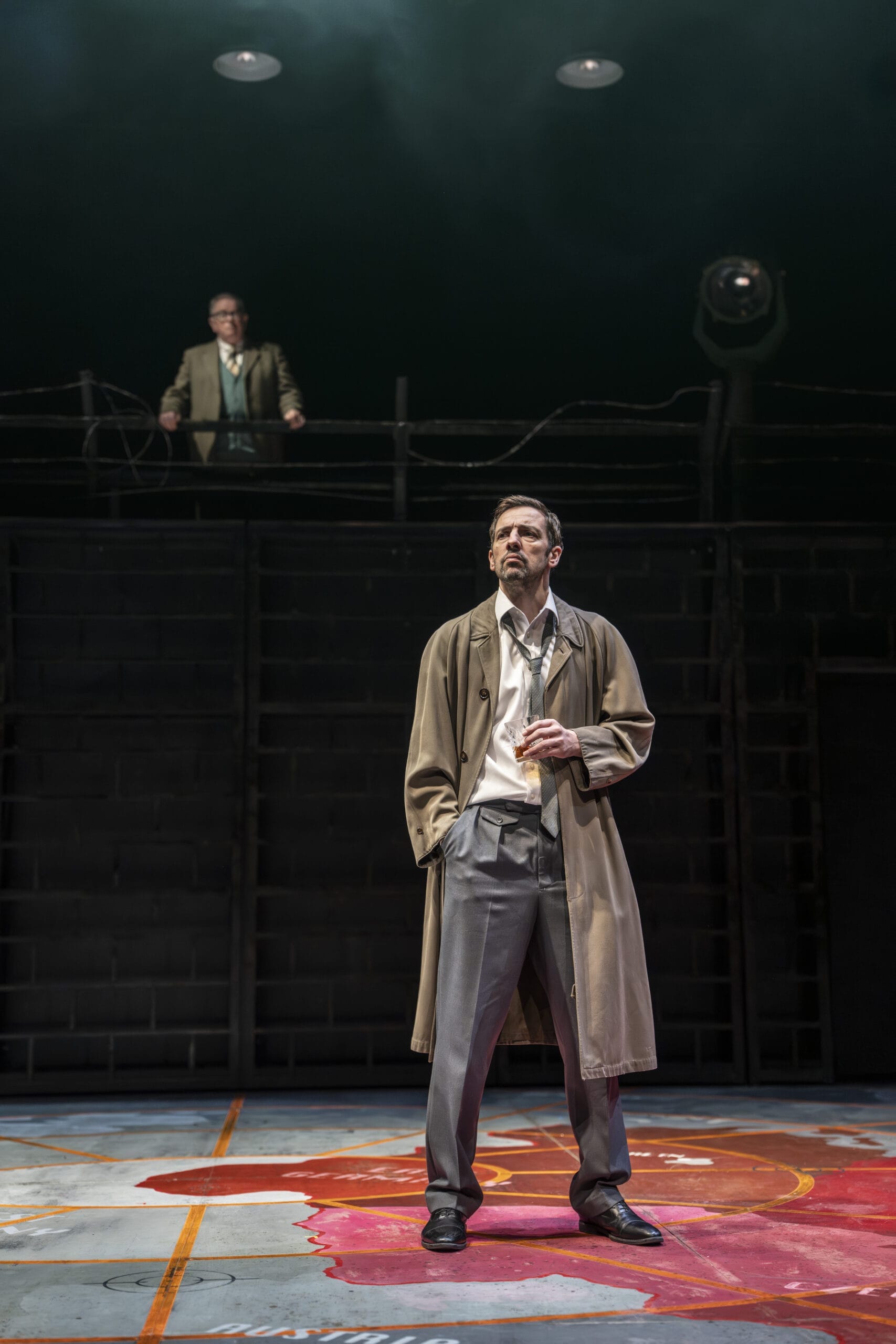 A man in a trench coat, evoking a John le Carré spy novel, stands on stage with one hand in his pocket and a cup in the other. Another man stands on a raised platform behind him. The dramatic lighting and industrial set heighten the intrigue.