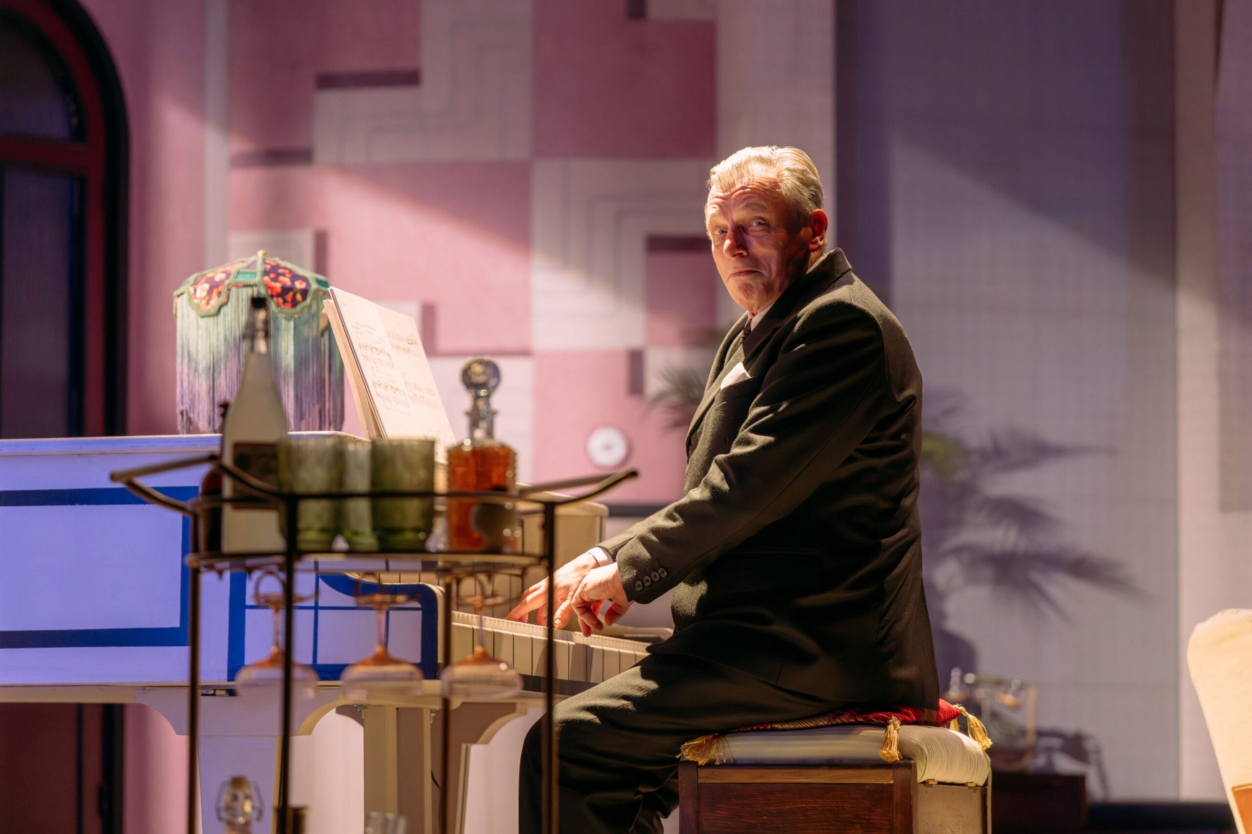A man in a suit sits at a white piano, looking toward the camera—sheet music open, drinks on a cart nearby—in a softly lit room reminiscent of "The Constant Wife.
