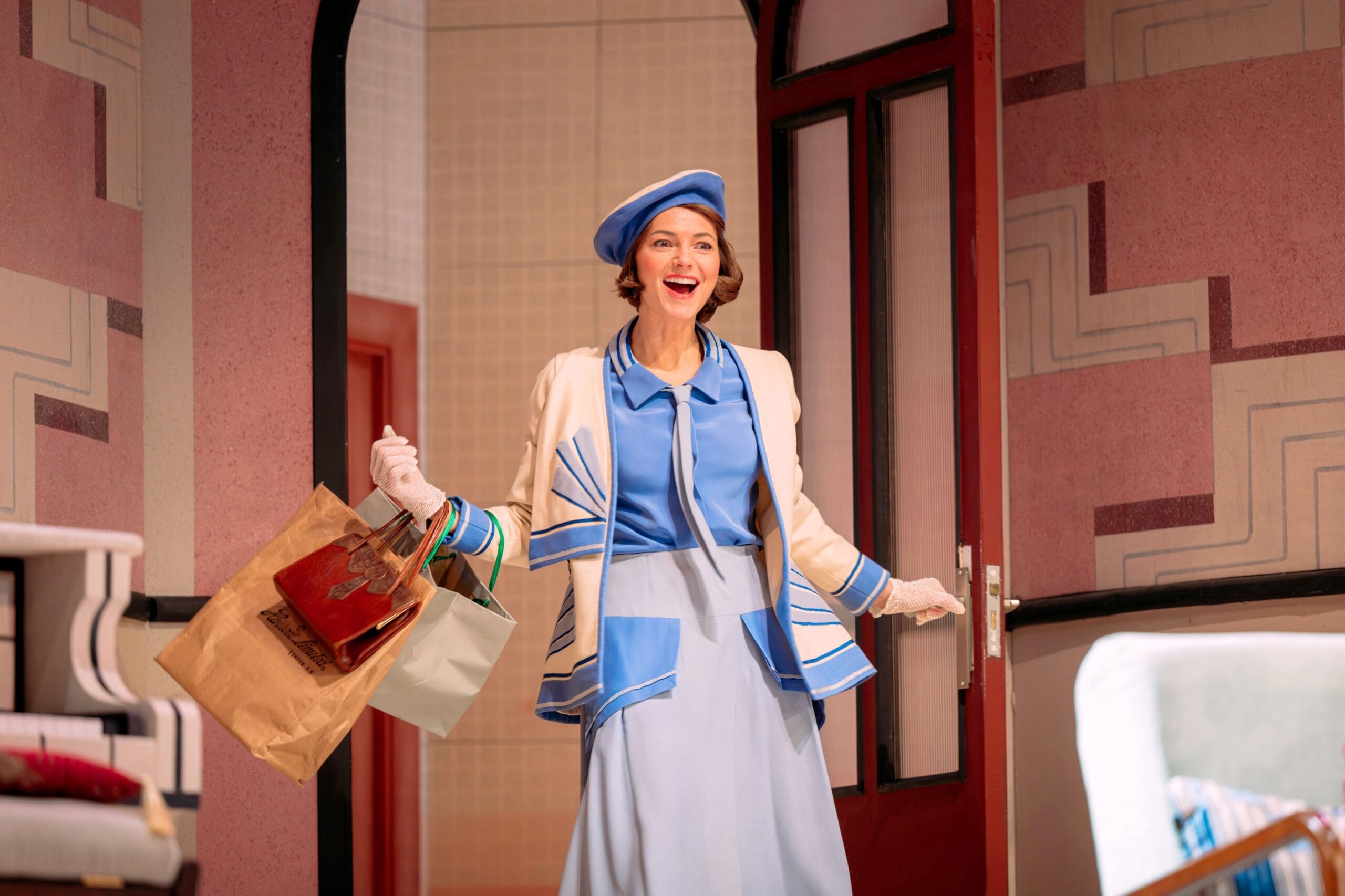 A woman in a blue and white outfit, reminiscent of The Constant Wife, enters a room smiling, holding shopping bags in both hands.