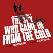 A man and a woman walk toward a fallen bicycle on a red background with bold text: "Spy Who Came in from the Cold," the gripping spy novel by John le Carré, adapted by David Eldridge and directed by Jeremy Herrin.