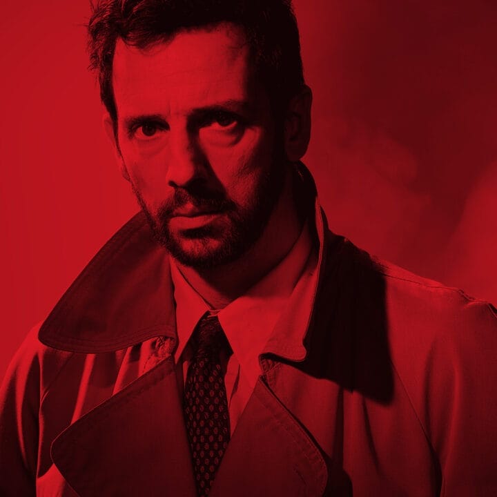 A serious man in a trench coat stands against a red background