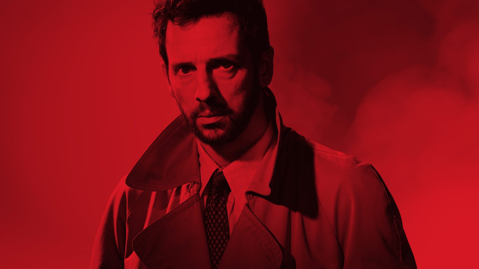 A serious man in a trench coat stands against a red background
