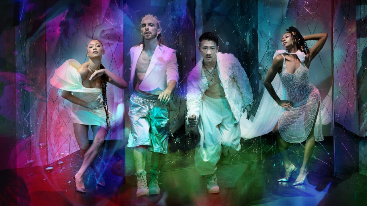 Four people pose in futuristic, metallic and sheer Rambert-inspired outfits against a vibrant, abstract, multicoloured background with blue, pink, and purple lighting effects.