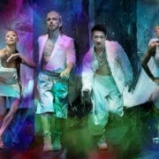 Four people pose in futuristic, metallic and sheer Rambert-inspired outfits against a vibrant, abstract, multicoloured background with blue, pink, and purple lighting effects.