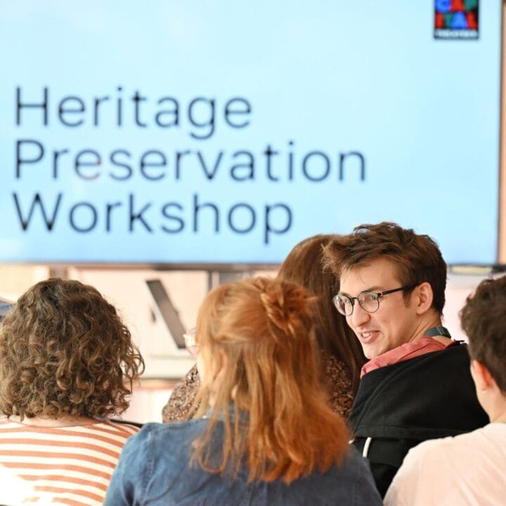People seated in a room face a large screen displaying the text "Heritage Preservation Workshop," where storytelling and preserving stories are at the heart of the event.