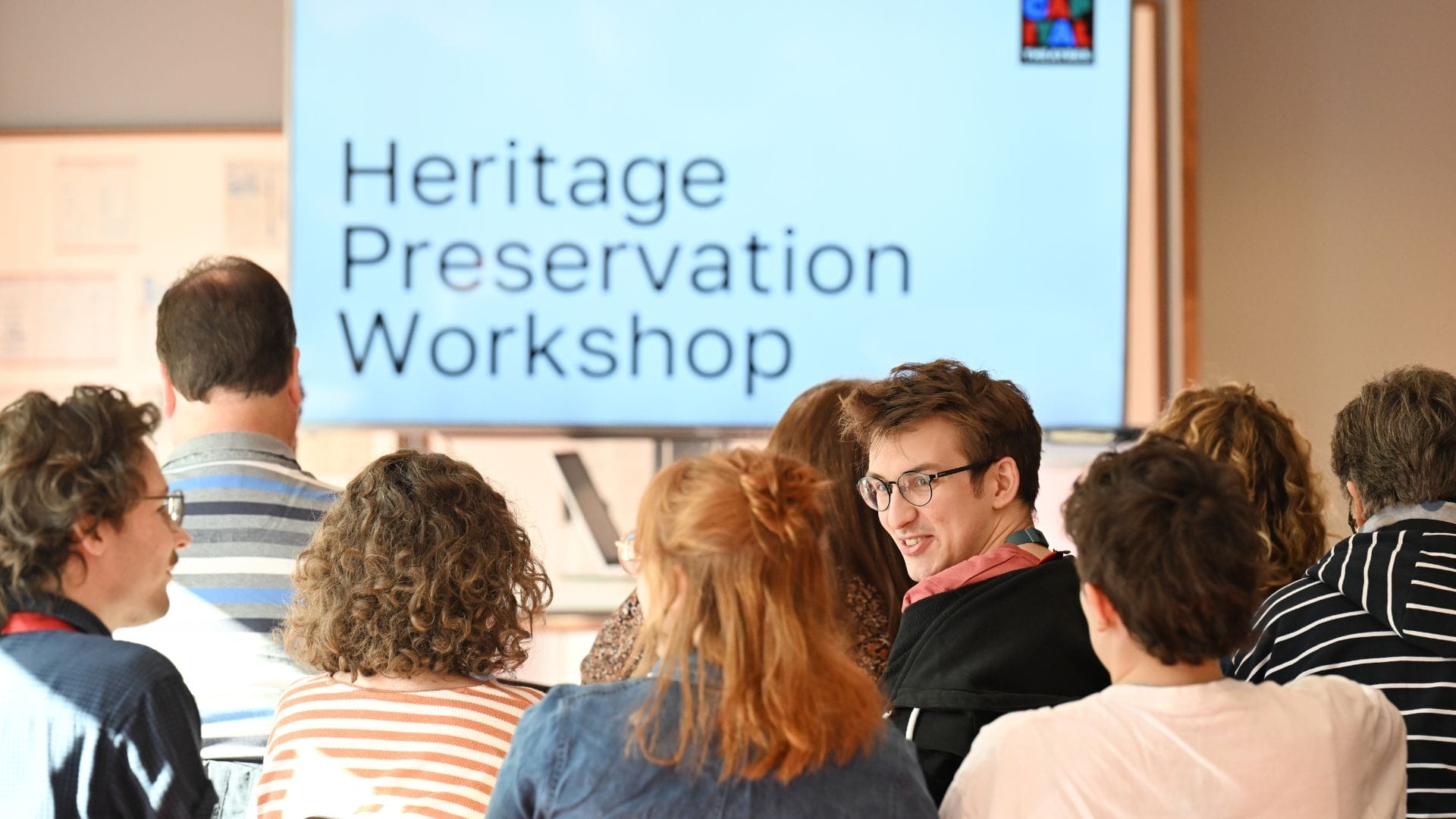 People seated in a room face a large screen displaying the text "Heritage Preservation Workshop," where storytelling and preserving stories are at the heart of the event.