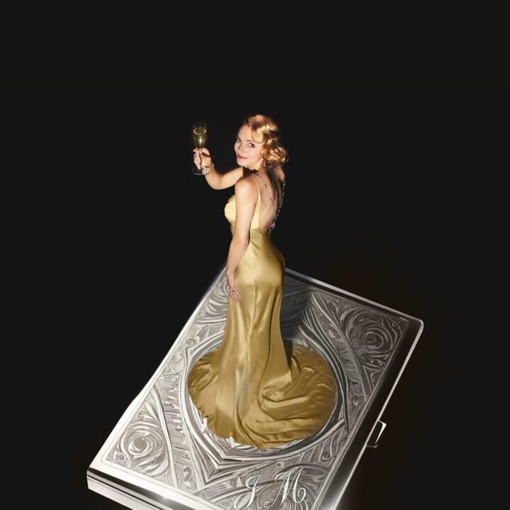 A woman in a gold dress, evoking a scene from The Constant Wife, holds a drink and stands on a large, ornate silver rectangular object against a black background.