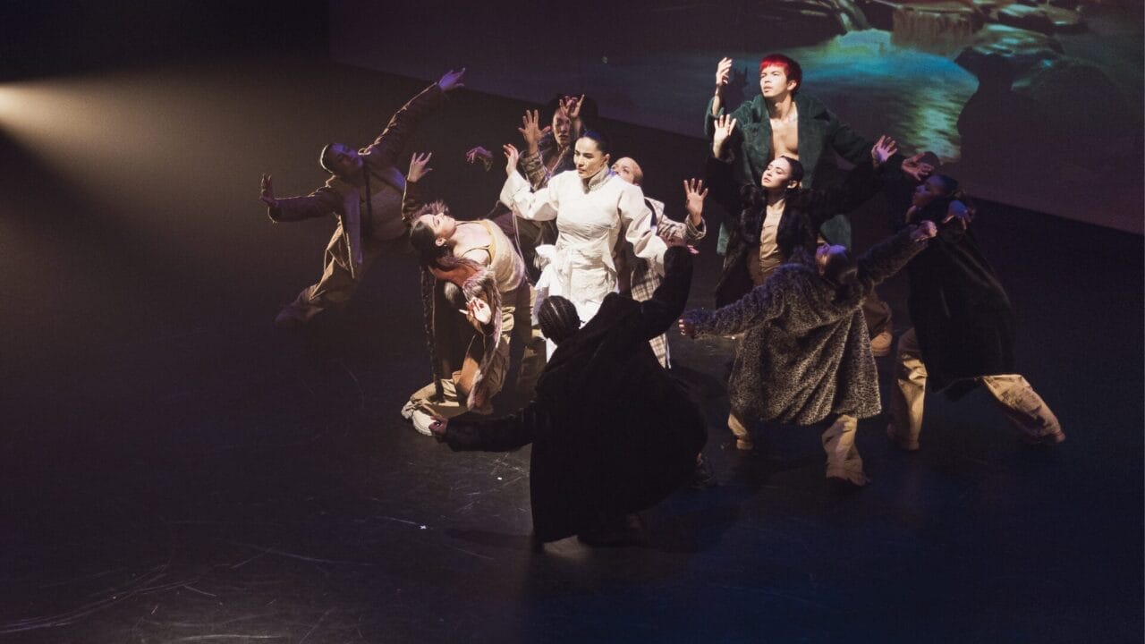 A group of dancers on stage, some in dark outfits and one in white, perform with arms raised under dramatic lighting, capturing the essence of Evolution in a striking Three60 World performance.