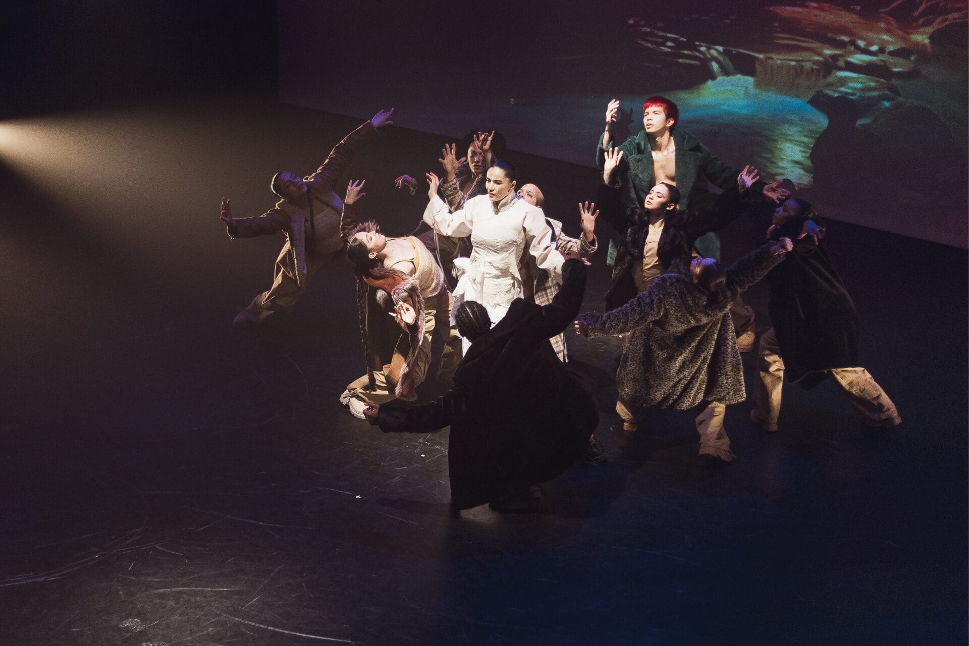 A group of dancers on stage, some in dark outfits and one in white, perform with arms raised under dramatic lighting, capturing the essence of Evolution in a striking Three60 World performance.