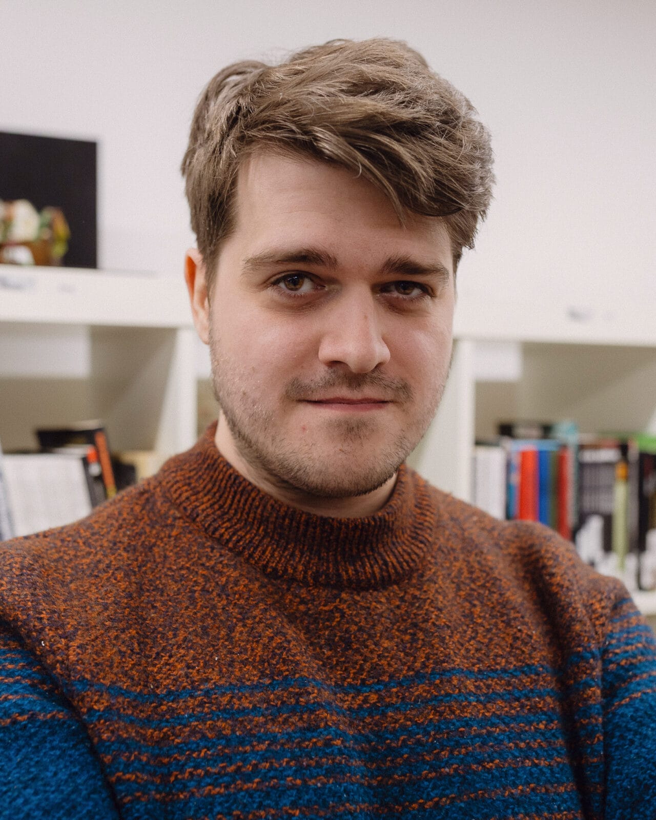 A man with short brown hair and a beard, wearing a brown and blue sweater, stands in front of bookshelves.