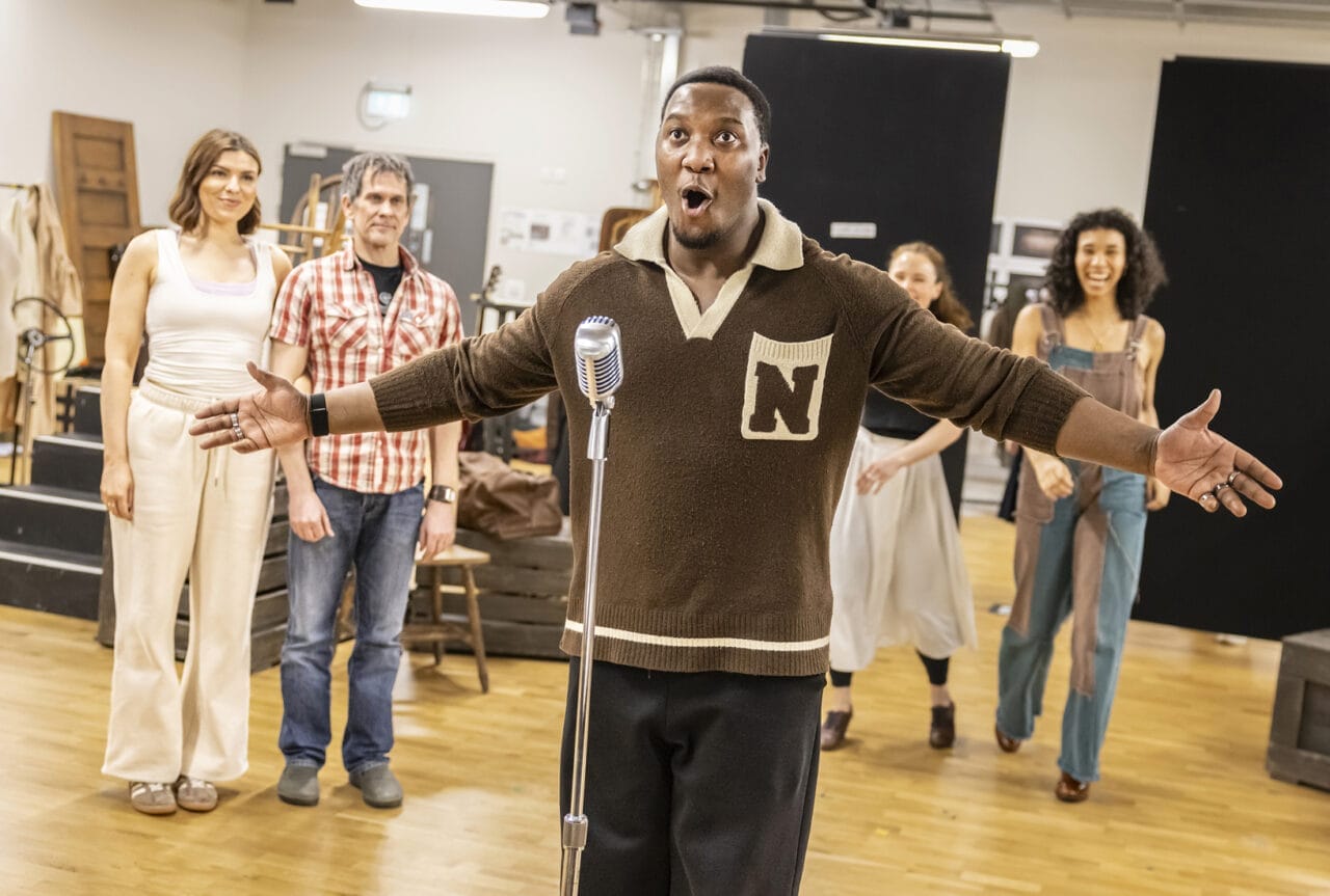 A man stands in front of a vintage-style microphone with his arms outstretched, appearing to sing a ballad, while four people watch him in the background of a rehearsal studio, evoking the spirit of classic music legends like Johnny and June.