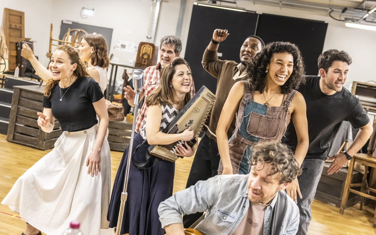 A group of seven adults in casual clothes and costumes are singing and playing instruments in a rehearsal room, with wooden chairs and props in the background, practising a ballad from Johnny and June for an upcoming musical or performance.