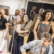 A group of seven adults in casual clothes and costumes are singing and playing instruments in a rehearsal room, with wooden chairs and props in the background, practising a ballad from Johnny and June for an upcoming musical or performance.