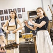 Two women perform indoors, one playing a flute and the other holding a washboard, evoking the spirit of a classic ballad. Wooden crates, stacked chairs, and a drum fill the background along with photographs on the wall.