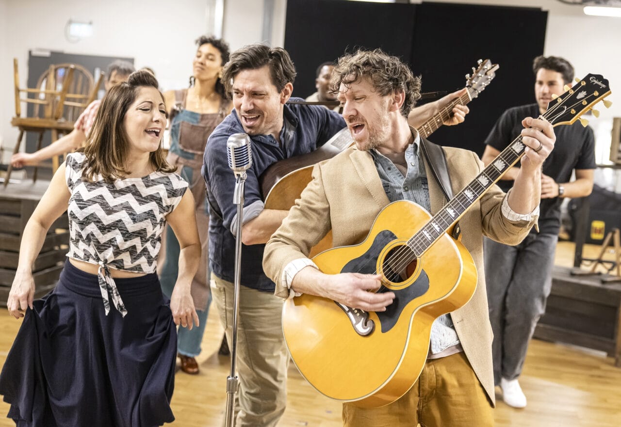 A group of musicians and singers perform enthusiastically indoors. Channelling Johnny and June, a man in the foreground plays an acoustic guitar beside a woman singing a heartfelt ballad into a retro-style microphone, whilst others dance and play instruments in the background.