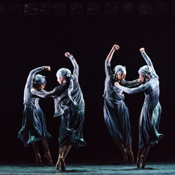 Four dancers in matching gray costumes and headscarves perform a synchronized ballet dance on a dimly lit stage, arms raised and legs extended, celebrating the Ballet Black anniversary.