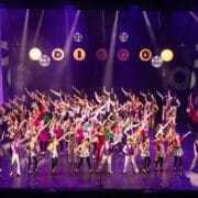 A large group of performers in red and white costumes are singing and dancing on a brightly lit theater stage with a cityscape backdrop at the 2026 Edinburgh Gang Show in Edinburgh.