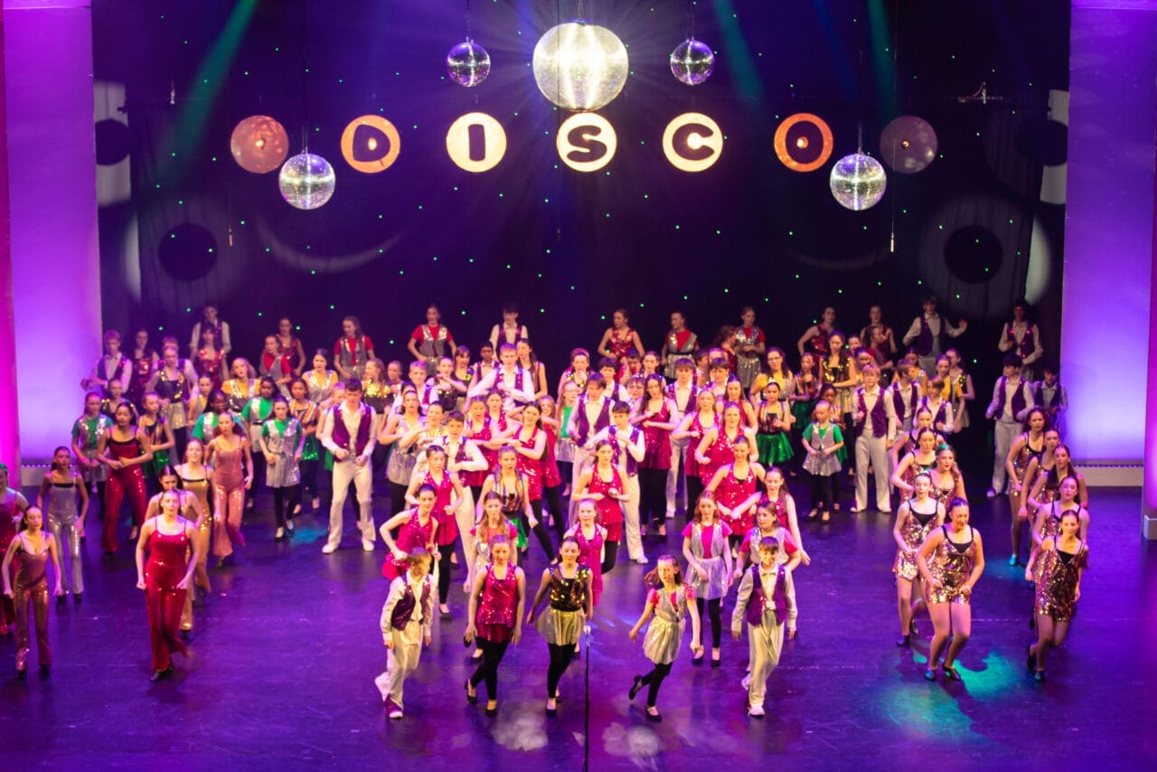A large group of performers in red and white costumes are singing and dancing on a brightly lit theater stage with a cityscape backdrop at the 2026 Edinburgh Gang Show in Edinburgh.