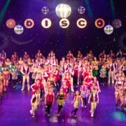 A large group of performers in red and white costumes are singing and dancing on a brightly lit theater stage with a cityscape backdrop at the 2026 Edinburgh Gang Show in Edinburgh.
