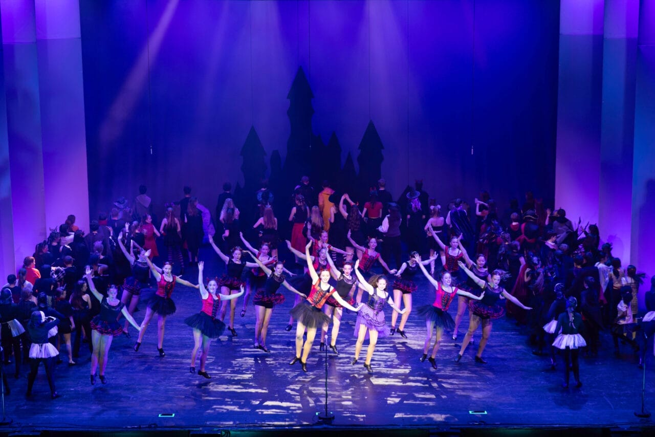 A large group of performers in red and white costumes are singing and dancing on a brightly lit theater stage with a cityscape backdrop at the 2026 Edinburgh Gang Show in Edinburgh.