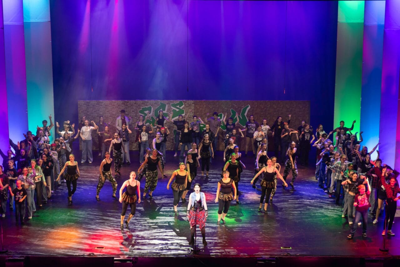 A large group of performers in red and white costumes are singing and dancing on a brightly lit theater stage with a cityscape backdrop at the 2026 Edinburgh Gang Show in Edinburgh.