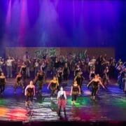 A large group of performers in red and white costumes are singing and dancing on a brightly lit theater stage with a cityscape backdrop at the 2026 Edinburgh Gang Show in Edinburgh.