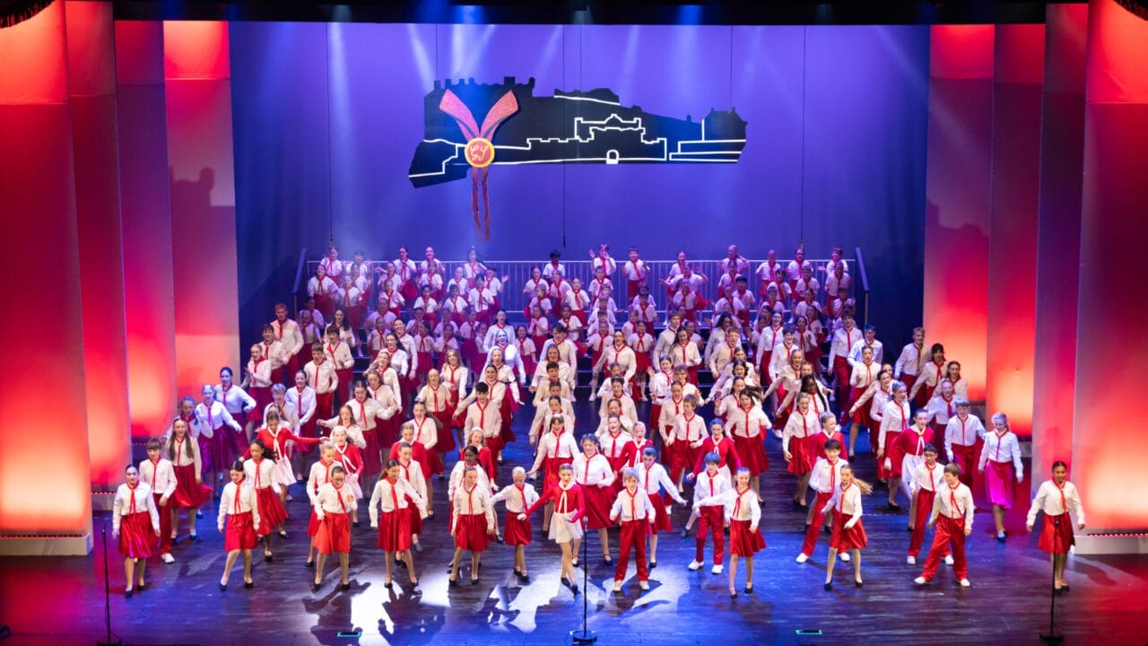 A large group of performers in red and white costumes are singing and dancing on a brightly lit theater stage with a cityscape backdrop at the 2026 Edinburgh Gang Show in Edinburgh.