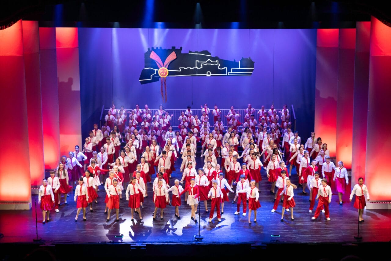 A large group of performers in red and white costumes are singing and dancing on a brightly lit theater stage with a cityscape backdrop at the 2026 Edinburgh Gang Show in Edinburgh.