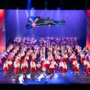 A large group of performers in red and white costumes are singing and dancing on a brightly lit theater stage with a cityscape backdrop at the 2026 Edinburgh Gang Show in Edinburgh.