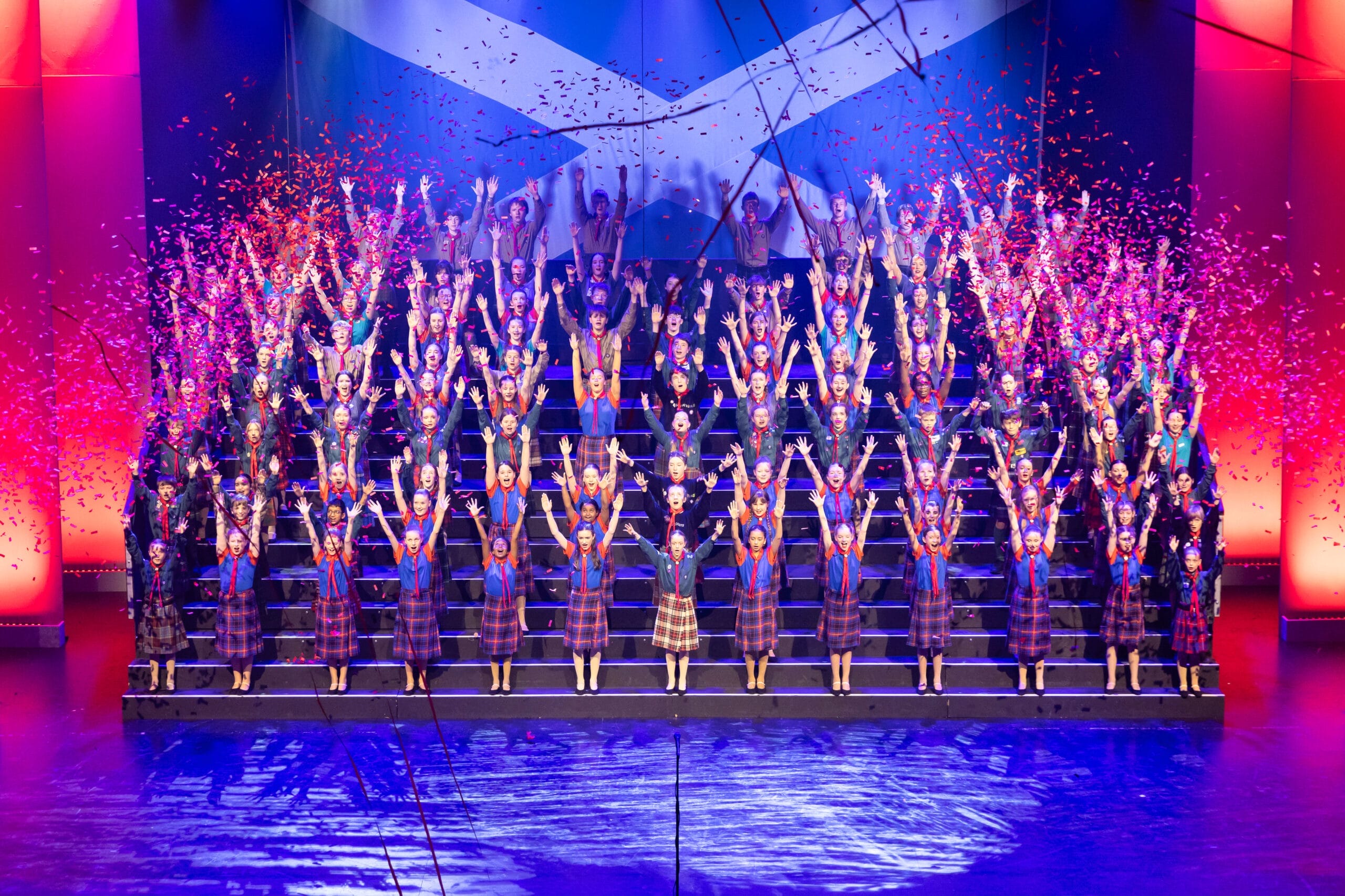 A large group of performers in red and white costumes are singing and dancing on a brightly lit theater stage with a cityscape backdrop at the 2026 Edinburgh Gang Show in Edinburgh.