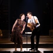 Two performers stand on stage, one singing into a microphone and the other playing guitar and singing, evoking a Johnny and June vibe as they deliver a heartfelt ballad under stage lighting.