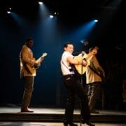 Man in white shirtsleeves singing on stage with big guitar