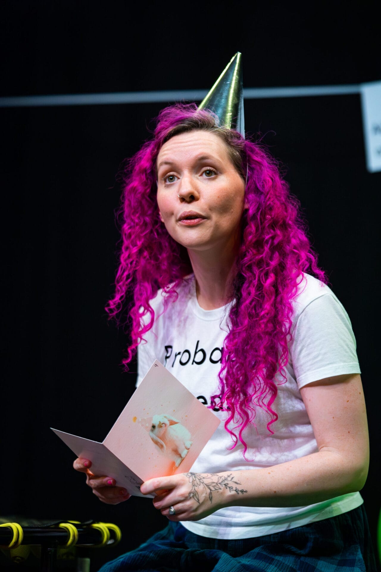 A person with bright pink curly hair, celebrating neurodiversity, wears a metallic party hat and holds an open greeting card while sitting against a dark background.