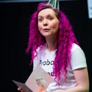 A person with bright pink curly hair, celebrating neurodiversity, wears a metallic party hat and holds an open greeting card while sitting against a dark background.