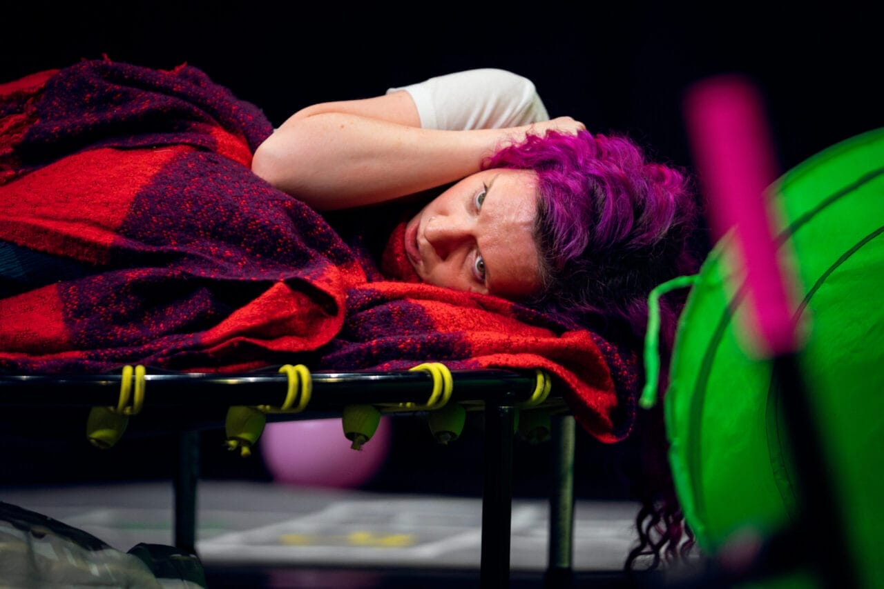 A person with vivid purple hair, embracing neurodivergence, lies on a red plaid blanket on a trampoline, resting their head on their arm and looking ahead. Bright green and pink objects are in the foreground.