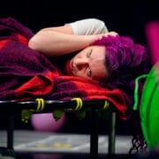 A person with vivid purple hair, embracing neurodivergence, lies on a red plaid blanket on a trampoline, resting their head on their arm and looking ahead. Bright green and pink objects are in the foreground.