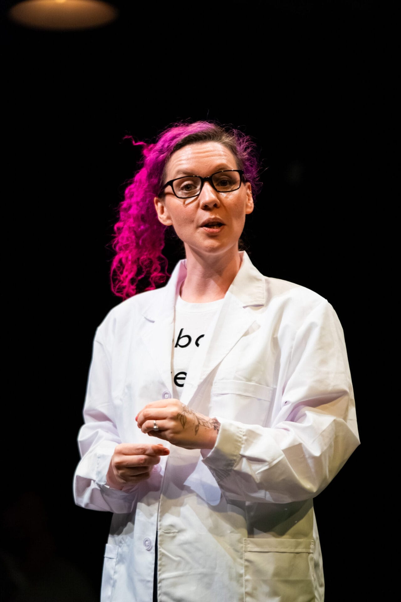 Person with bright pink hair and glasses wearing a white lab coat, standing and speaking against a dark background, passionately discussing neurodiversity in science.