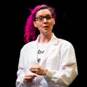 Person with bright pink hair and glasses wearing a white lab coat, standing and speaking against a dark background, passionately discussing neurodiversity in science.