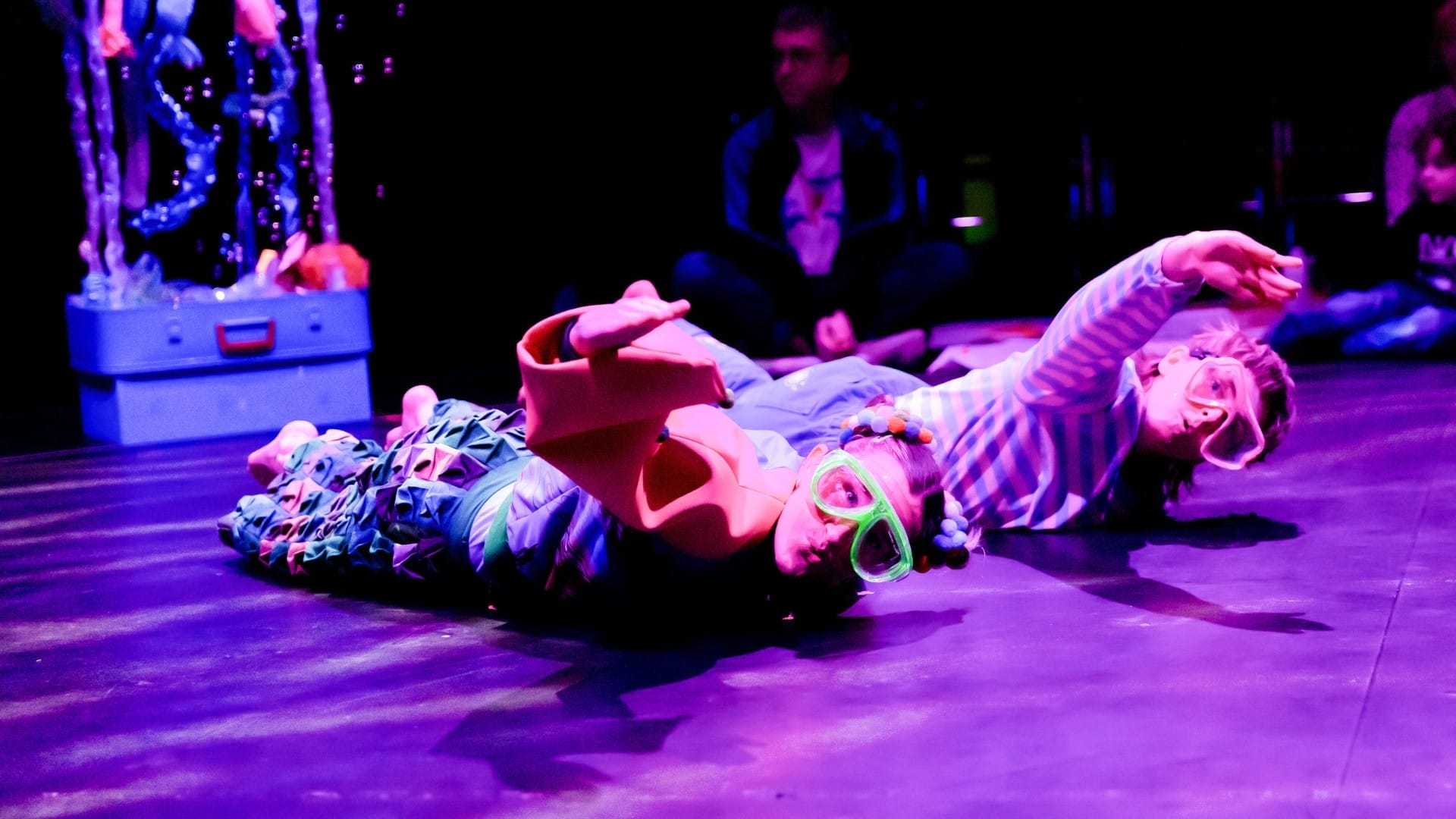 Two children wearing goggles lie on the floor under purple lighting, with one adult—perhaps from Home For Hamish—seated in the background.