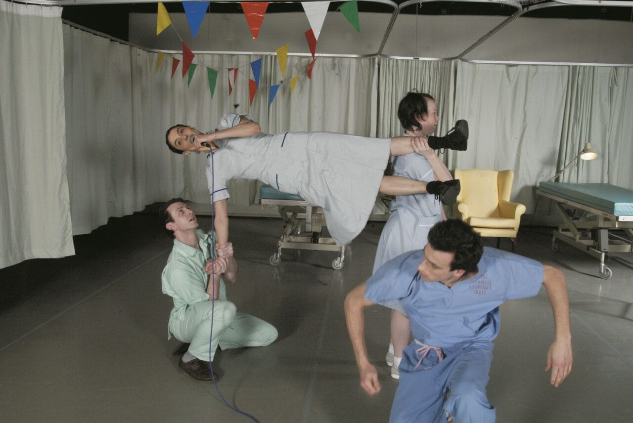 Four people in medical uniforms perform a playful, acrobatic pose in a hospital room decorated with colorful pennant banners.