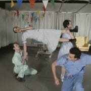 Four people in medical uniforms perform a playful, acrobatic pose in a hospital room decorated with colorful pennant banners.